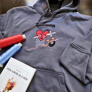 K&ouml;nnte beinhalten: Ein grauer Kapuzenpullover mit einem roten Herzen mit einer Tr&auml;ne und einem Messer, das durchsto&szlig;en wird, einer Schlange mit roten und wei&szlig;en Streifen, einer brennenden Acht und dem Text "POR AMOR AL CAOS" auf einem Buch im Vordergrund.