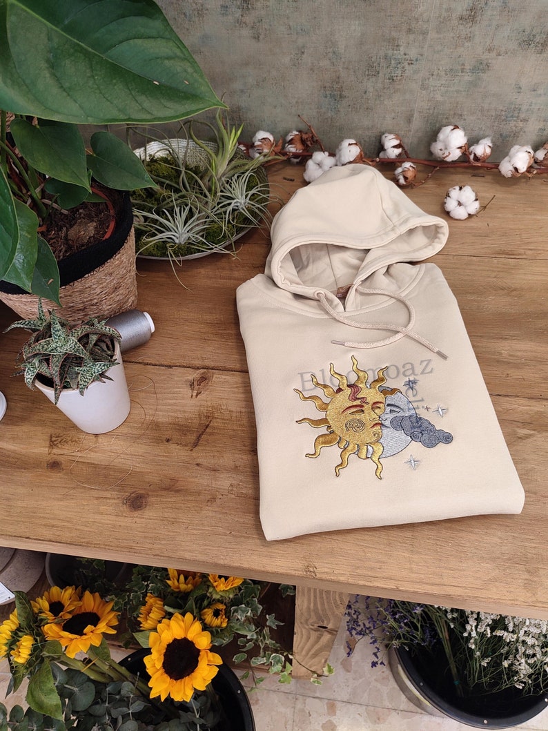 May include: A beige hoodie with a sun and moon design embroidered on the front. The design features a sun with a face and a crescent moon with a cloud. The text "El Sol y La Paz" is embroidered above the design.