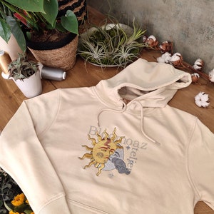 May include: A cream colored hoodie with a sun and moon design embroidered on the front. The design features a sun with a face and a crescent moon with a face. The text "Bloom" and "atelier" is embroidered on the hoodie.
