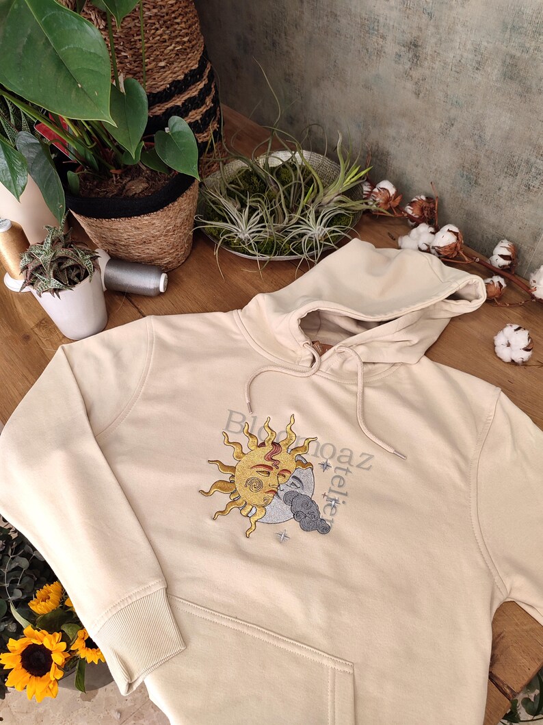 May include: A cream colored hoodie with a sun and moon design embroidered on the front. The design features a sun with a face and a crescent moon with a face. The text "Bloom" and "atelier" is embroidered on the hoodie.
