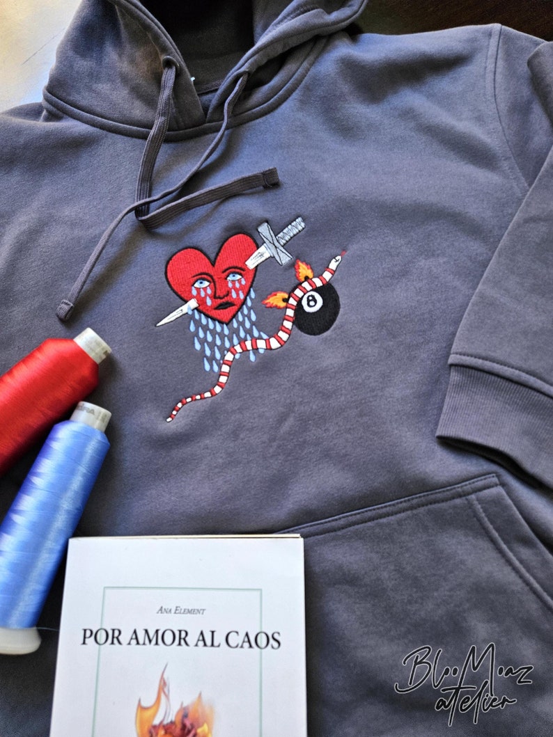 K&ouml;nnte beinhalten: Grauer Kapuzenpullover mit einem gestickten Design eines roten Herzens mit einer Tr&auml;ne, einem Dolch, einer Schlange und einer schwarzen 8-Ball. Der Text "POR AMOR AL CAOS" ist auf einem Buch im Bild gedruckt.