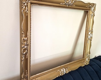 Könnte beinhalten: Verzierter goldener Bilderrahmen mit detaillierten floralen und scrollartigen Verzierungen. Der rechteckige Rahmen hat einen gewellten oberen Rand und silberne Akzente. Er lehnt an einer weißen Wand und einer blauen Oberfläche.