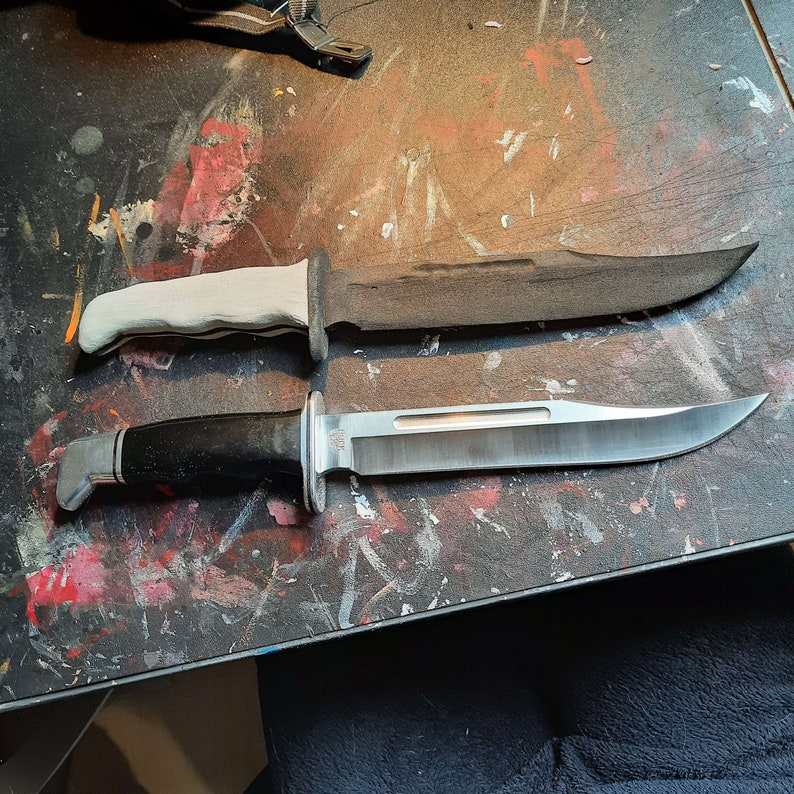 May include: Two hunting knives with fixed blades. One knife has a black handle and a silver blade, while the other has a white handle and a dark blade. Both knives are on a multi-colored surface.