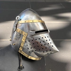 Pu&ograve; includere: Un elmo medievale color argento con dettagli in ottone e rivetti. L'elmo presenta una visiera appuntita con piccoli fori e una stretta fessura per gli occhi. Sono attaccati una catena e un cinturino in pelle. Il testo "KNIGHTCRAFT HOUSE" &egrave; visibile.