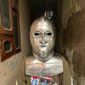 Op de afbeelding: Een zilveren ridderhelm met een gezichtsbeschermer en een nekbeschermer. De helm wordt gedragen door een persoon die een groen T-shirt draagt met een grafische print van een cartoonfiguur. De tekst "KnightCraftHouse" is zichtbaar op het T-shirt.
