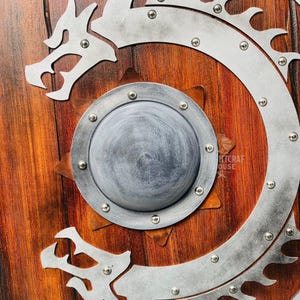 May include: A wooden shield with a silver dragon design. The shield has a round, silver center with a raised, starburst pattern. The dragon is curled around the shield, with its head facing the viewer. The shield is decorated with silver rivets.