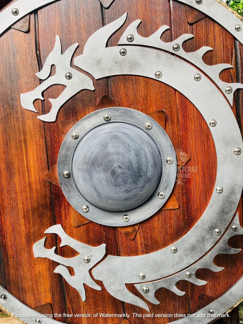 May include: A wooden shield with a silver dragon design. The shield has a round, silver center with a raised, starburst pattern. The dragon is curled around the shield, with its head facing the viewer. The shield is decorated with silver rivets.