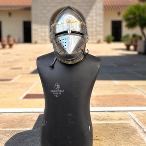 Pu&ograve; includere: Un elmo da cavaliere in acciaio lucido con dettagli in ottone, esposto su un manichino nero. L'elmo presenta una visiera con fessure per gli occhi e numerosi fori di ventilazione. Il manichino ha il testo "KNIGHTCRAF THOUSE" stampato.