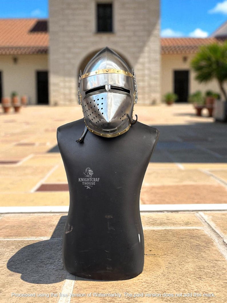 Pu&ograve; includere: Un elmo da cavaliere in acciaio lucido con dettagli in ottone, esposto su un manichino nero. L'elmo presenta una visiera con fessure per gli occhi e numerosi fori di ventilazione. Il manichino ha il testo "KNIGHTCRAF THOUSE" stampato.