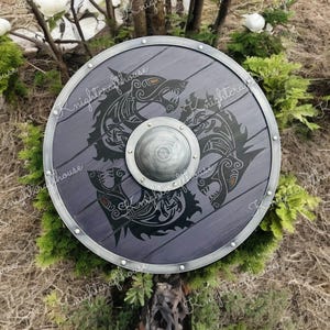 Puede incluir: Un escudo redondo de madera con un borde de metal y un diseño en negro y gris de dos lobos luchando. El escudo está decorado con remaches alrededor del borde.