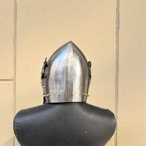 Pu&ograve; includere: Un elmo medievale argentato lucido con la parte superiore a punta, esposto su un manichino nero. L'elmo presenta elementi decorativi e il testo "KNIGHTCRAFT THONSE".