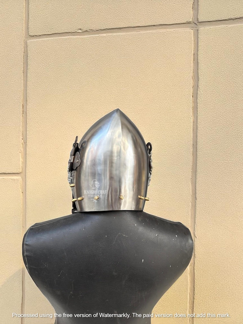 Pu&ograve; includere: Un elmo medievale argentato lucido con la parte superiore a punta, esposto su un manichino nero. L'elmo presenta elementi decorativi e il testo "KNIGHTCRAFT THONSE".