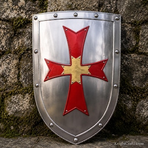 Könnte beinhalten: Ein polierter silberner Wappenschild mit einem markanten roten und goldenen Kreuzemblem in der Mitte. Der Schild ist von einem erhöhten Metallrand umgeben, der mit gleichmäßig verteilten Nieten befestigt ist. Der Text KnightCraftHouse ist unten sichtbar.