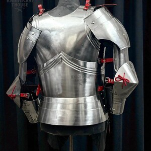 May include: Polished steel armor set, featuring a breastplate, shoulder guards, and arm protection. Red leather straps and accents secure the pieces. The armor is displayed on a mannequin. The text "KNIGHTCRAFT HOUSE" is visible.