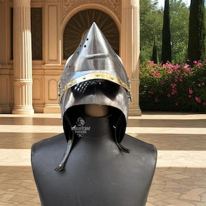 Pu&ograve; includere: Un elmo da cavaliere in metallo lucido con una parte superiore a punta e dettagli in ottone. L'elmo &egrave; esposto su una testa di manichino nera. Il testo "KNIGHTCRAF THOUSE" &egrave; visibile sul collo del manichino.
