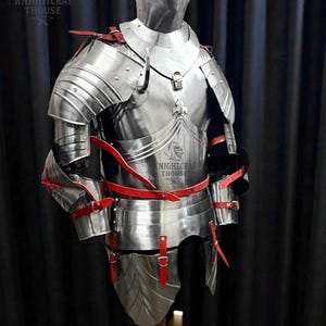 May include: A full suit of polished steel armor with red leather straps and accents. The armor includes a helmet, chest plate, shoulder guards, and leg protection. The Knightcrafthouse logo is visible.