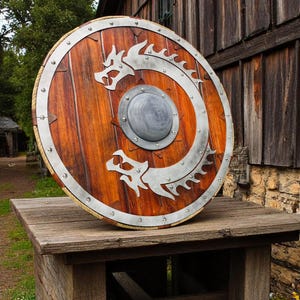 May include: A round wooden shield with a metal rim and a silver dragon design. The shield is resting on a wooden table.