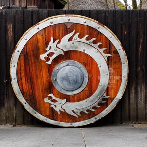 May include: A round wooden shield with a metal rim and a silver dragon design. The shield has a metal boss in the center and is decorated with metal rivets.