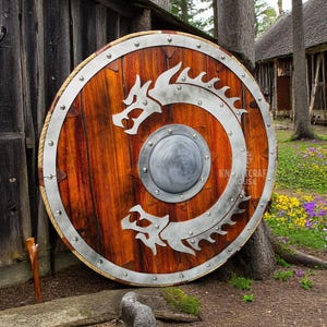May include: A large, round wooden shield with a metal rim and two dragon designs. The shield is decorated with metal rivets and has a smaller metal circle in the center.