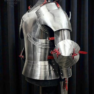 May include: A polished steel breastplate and arm armor with red leather straps and accents. The armor is displayed on a mannequin, showcasing its intricate design and craftsmanship. The image includes the text "KNIGHTCRAFT HOUSE".
