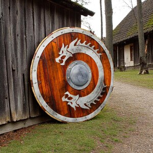 May include: A large, round wooden shield with a metal rim and two silver dragons intertwined on the front. The shield is leaning against a wooden wall.