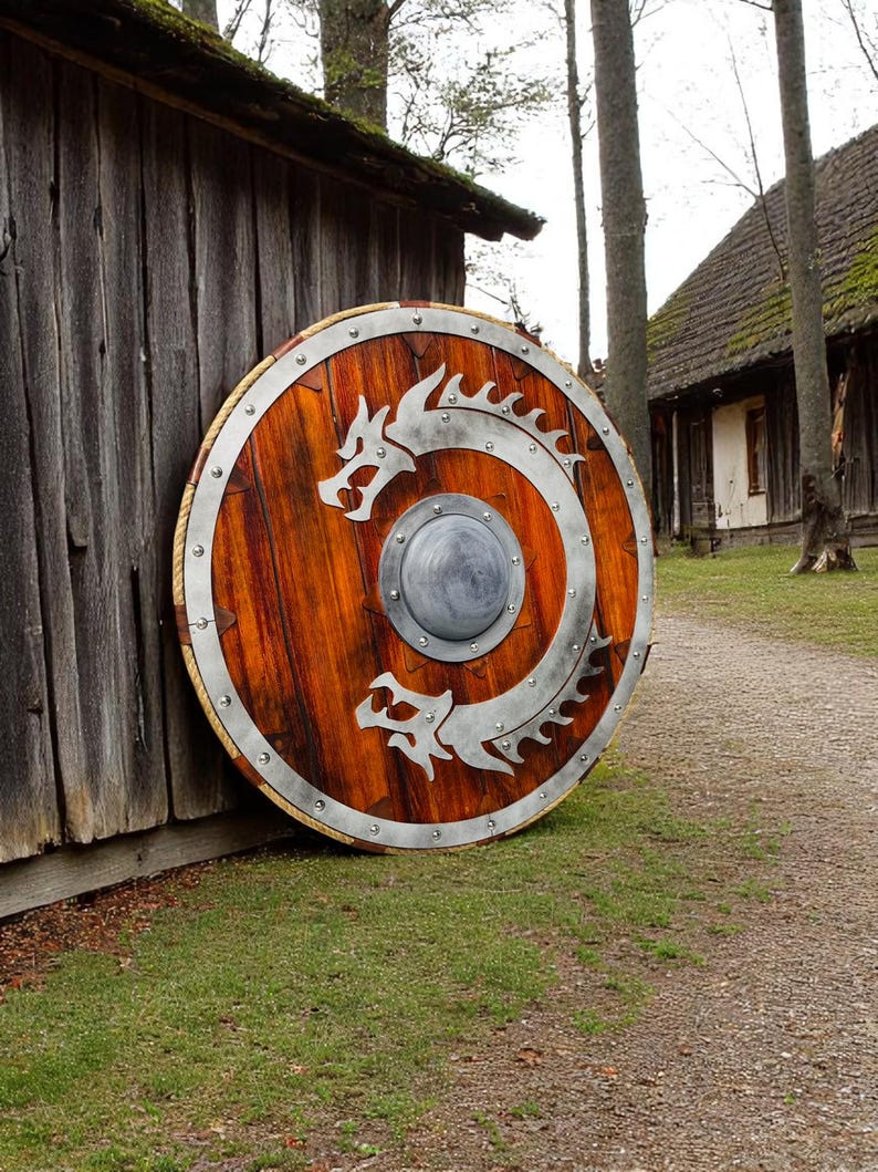 May include: A large, round wooden shield with a metal rim and two silver dragons intertwined on the front. The shield is leaning against a wooden wall.