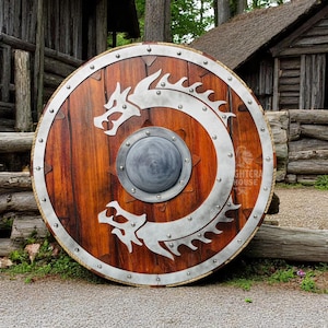May include: A round wooden shield with a silver dragon design. The shield has a metal rim with rivets and a small metal boss in the center. The shield is leaning against a wooden building.
