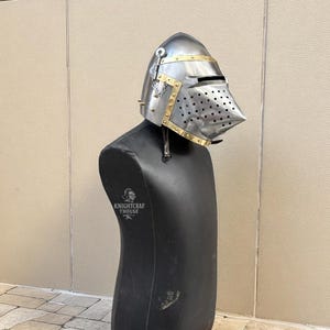 Pu&ograve; includere: Un elmo in stile medievale in metallo argentato con dettagli in ottone. L'elmo &egrave; esposto su un manichino nero. Il testo "KNIGHTCRAF THOUSE" &egrave; visibile sul manichino.