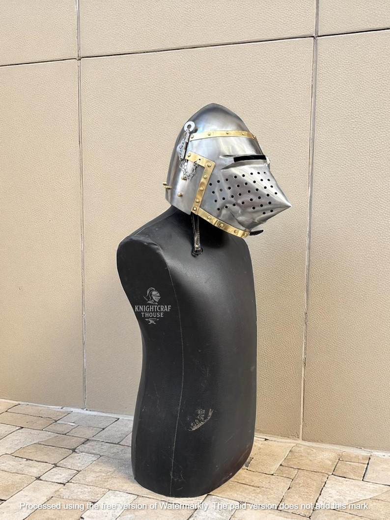 Pu&ograve; includere: Un elmo in stile medievale in metallo argentato con dettagli in ottone. L'elmo &egrave; esposto su un manichino nero. Il testo "KNIGHTCRAF THOUSE" &egrave; visibile sul manichino.