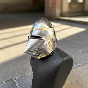 Pu&ograve; includere: Un elmo in acciaio lucido con visiera a punta e dettagli in ottone. L'elmo &egrave; esposto su una testa di manichino nera. Presenta una catena e un piccolo stemma. Il testo "KNIGHTCRAF THOUSE" &egrave; visibile.