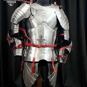May include: A full suit of polished steel armor with red leather straps and accents. The armor is displayed on a mannequin, showcasing its intricate design and craftsmanship. The Knightcrafthouse logo is visible.