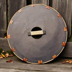 May include: A round, dark brown shield with a rope handle and brass rivets around the edge. The shield is leaning against a wooden fence.