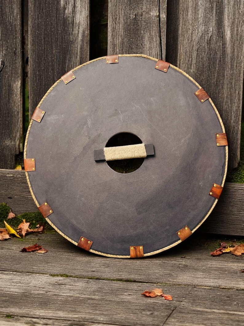 May include: A round, dark brown shield with a rope handle and brass rivets around the edge. The shield is leaning against a wooden fence.