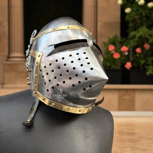Pu&ograve; includere: Un elmo medievale color argento con dettagli in ottone. L'elmo presenta una visiera appuntita, numerosi fori di ventilazione e il testo "KNIGHTCRAF THOUSE". &Egrave; esposto su una superficie nera.