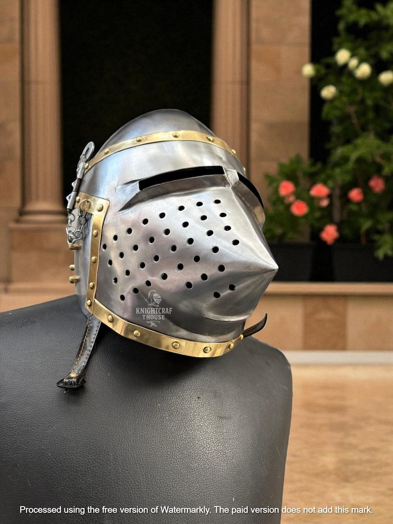 Pu&ograve; includere: Un elmo medievale color argento con dettagli in ottone. L'elmo presenta una visiera appuntita, numerosi fori di ventilazione e il testo "KNIGHTCRAF THOUSE". &Egrave; esposto su una superficie nera.