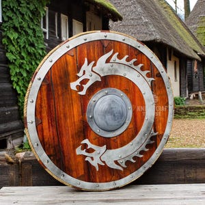 May include: A round wooden shield with a silver metal rim and two silver dragons on the front. The shield has a small round metal boss in the center. The shield is sitting on a wooden table.