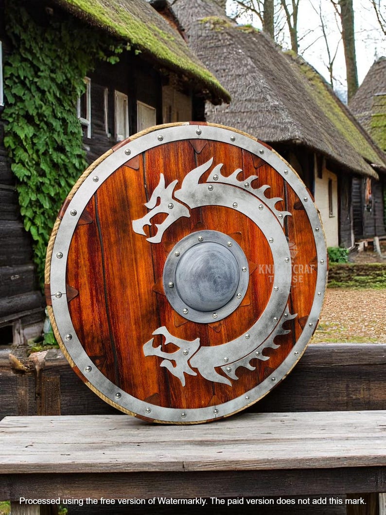 May include: A round wooden shield with a silver metal rim and two silver dragons on the front. The shield has a small round metal boss in the center. The shield is sitting on a wooden table.