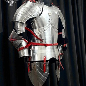 May include: A full suit of polished steel armor with red leather straps and accents. The armor is displayed on a mannequin, showcasing its intricate design and craftsmanship. The image includes the text "KNIGHTCRAF THOUSE".