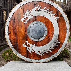 May include: A round wooden shield with a metal rim and a silver dragon design. The shield has a small metal boss in the center.
