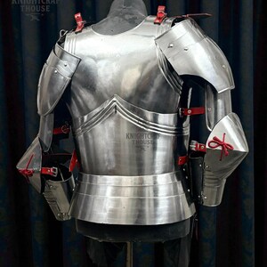 May include: A polished steel medieval armor breastplate and shoulder guards, with red leather straps and accents. The armor is displayed on a mannequin, showcasing its intricate design and craftsmanship. The Knightcrafthouse logo is visible.