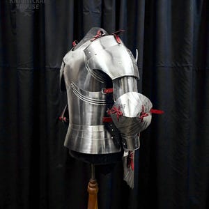 May include: A polished steel breastplate and arm armor with red leather accents. The armor is displayed on a mannequin against a black backdrop. The image includes the text "KNIGHTCRAFT HOUSE".