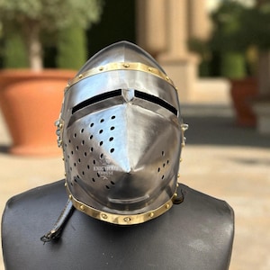 Pu&ograve; includere: Un elmo medievale color argento con dettagli in ottone e piccoli fori di ventilazione. L'elmo presenta una visiera con fessure per gli occhi ed &egrave; esposto su una testa di manichino nera. Il testo "KNIGHTCRAFT HOUSE" &egrave; visibile.