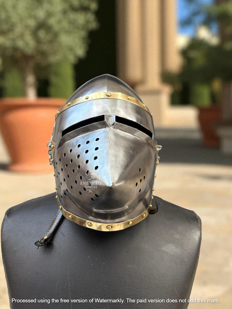 Pu&ograve; includere: Un elmo medievale color argento con dettagli in ottone e piccoli fori di ventilazione. L'elmo presenta una visiera con fessure per gli occhi ed &egrave; esposto su una testa di manichino nera. Il testo "KNIGHTCRAFT HOUSE" &egrave; visibile.