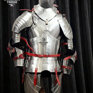 May include: A full suit of polished steel armor with red leather straps and accents. The armor includes a helmet, chest plate, shoulder guards, and leg protection. The armor is displayed on a wooden stand against a dark background. The Knightcrafthouse logo is visible.