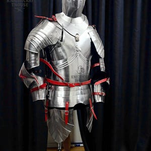 May include: A full suit of polished steel armor with red leather straps and accents. The armor includes a helmet, chest plate, shoulder guards, and leg protection. The armor is displayed on a wooden stand against a dark curtain.