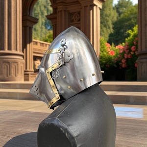 Pu&ograve; includere: Un elmo medievale in acciaio lucido con dettagli in ottone e rivetti. L'elmo presenta una parte superiore appuntita, una visiera con fori di ventilazione e una catena. &Egrave; esposto su un manichino nero, mostrando il suo design storico.