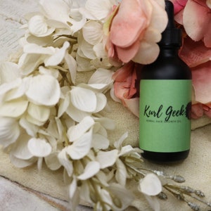 May include: A green glass bottle with a black dropper top and a green label that reads "Kurl Geek's Herbal Hair Growth Oil". The bottle is sitting on a white surface with pink and white flowers.