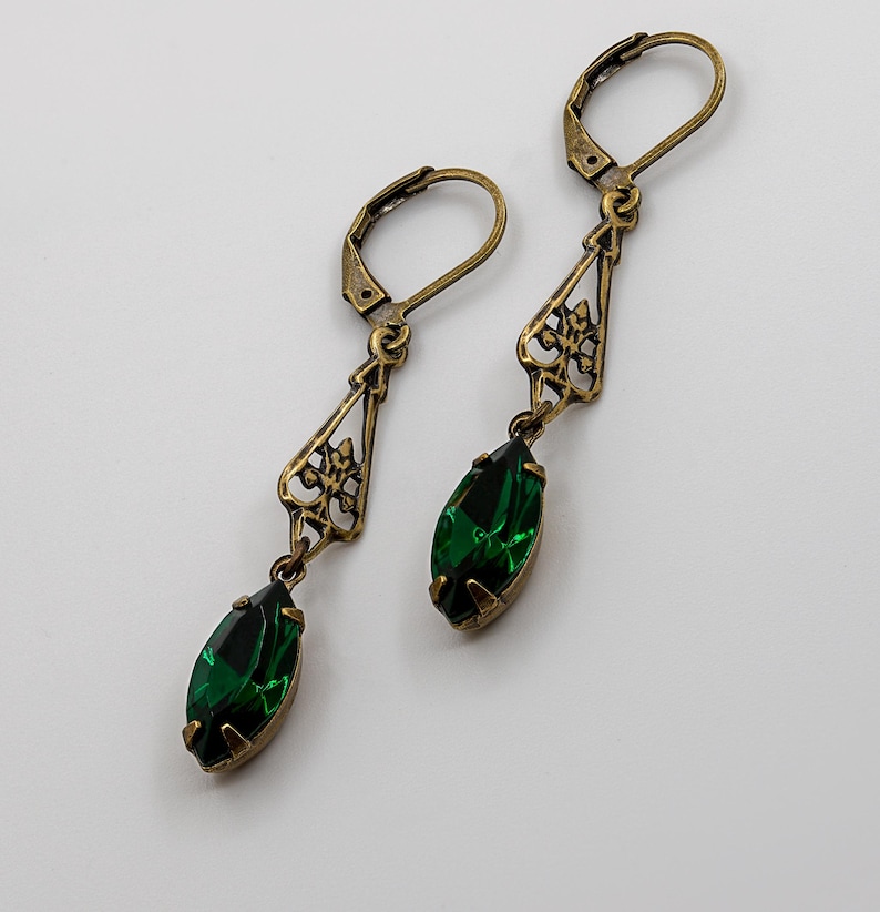 Pu&ograve; includere: Un paio di orecchini in stile antico con finitura bronzo. Ogni orecchino presenta un design a filigrana decorativo e una gemma verde smeraldo a forma di goccia. Gli orecchini hanno una chiusura a leva.