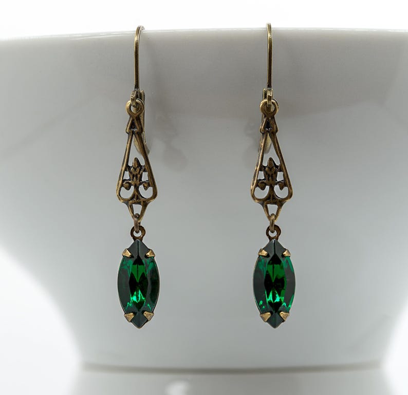 Pu&ograve; includere: Un paio di orecchini color oro antico. Ogni orecchino presenta un design a filigrana con un pendente a forma di marquise in cristallo verde smeraldo. Gli orecchini sono sospesi a un semplice gancio.