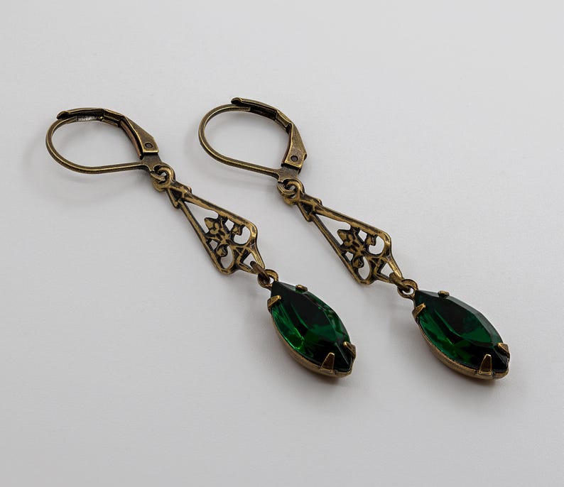 Pu&ograve; includere: Un paio di orecchini in stile antico con finitura bronzo. Ogni orecchino presenta un design a filigrana decorativo e una gemma verde smeraldo a forma di goccia. Gli orecchini hanno una chiusura a leva.